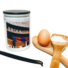 Load image into Gallery viewer, KCMOCO. Brookside Boulevard Candle in KC Collection Jar: White glass jar with photo label depicting Brookside Shops awning, with black bamboo lid. Egg, vanilla beans, wooden spoons and pile of flour in foreground.

