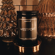 Load image into Gallery viewer, KCMOCO. candles Westport Candle in Classic Collection Jar: Black glass jar with gold foil label reading &quot;KCMOCO. Westport&quot;, with black bamboo lid, environmental photo  against backdrop of gold Christmas trees.
