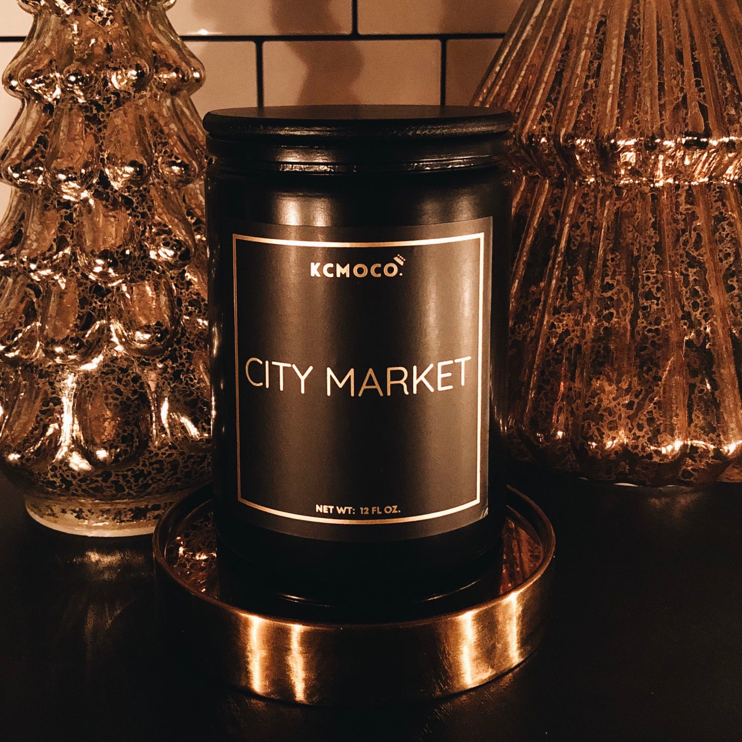 KCMOCO. Candles City Market Candle in Classic Collection Jar: Black glass jar with gold foil label reading "KCMOCO. City Market", with black bamboo lid, environmental photo against backdrop of gold Christmas trees.