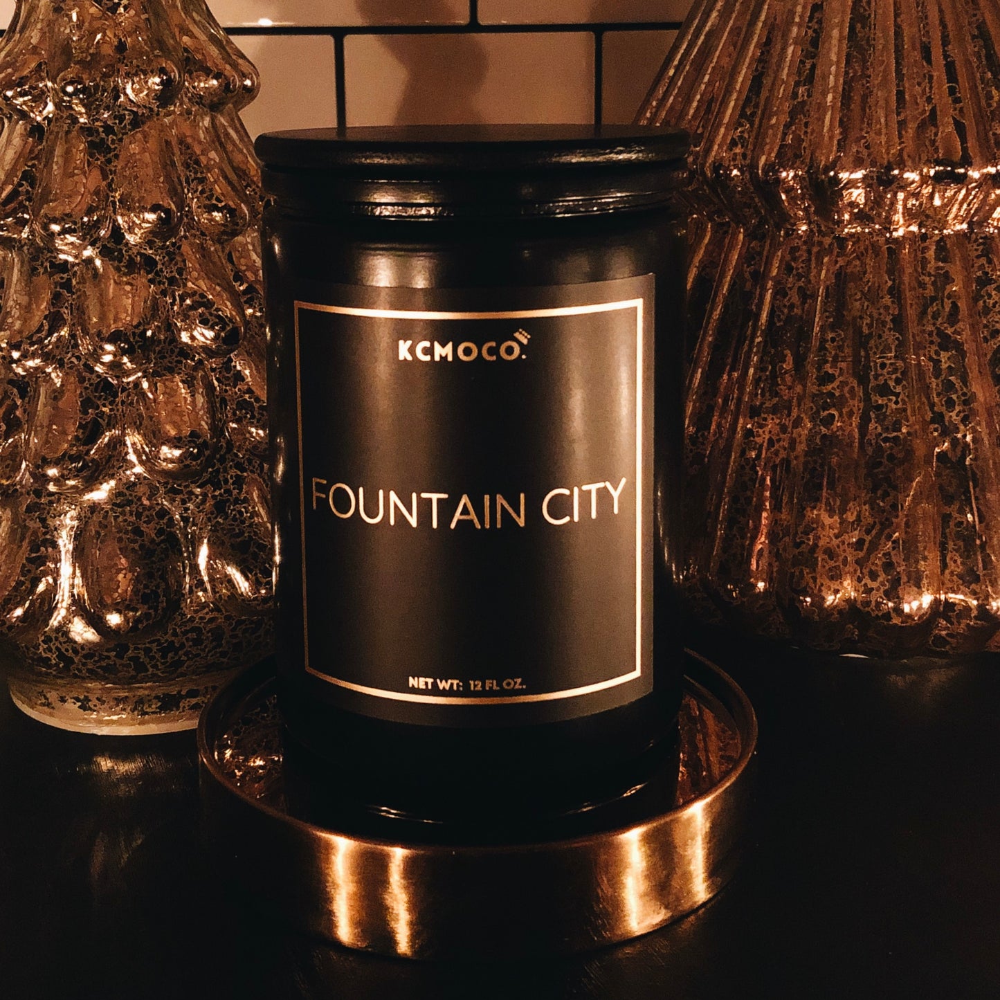 KCMOCO. candles City Market Candle in Classic Collection Jar: Black glass jar with gold foil label reading "KCMOCO. Fountain City", with black bamboo lid, environmental photo  against backdrop of gold Christmas trees.