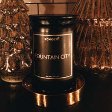 Load image into Gallery viewer, KCMOCO. candles City Market Candle in Classic Collection Jar: Black glass jar with gold foil label reading &quot;KCMOCO. Fountain City&quot;, with black bamboo lid, environmental photo  against backdrop of gold Christmas trees.
